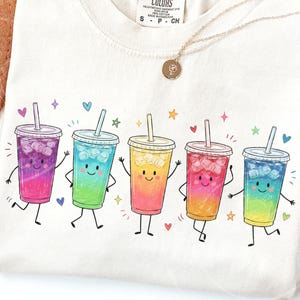 Puede incluir: Una camiseta de color crema con un gráfico de cinco vasos de bebida helada antropomórficos con líquidos de colores degradados, cubitos de hielo y pajitas. Cada vaso tiene una cara sonriente, brazos y piernas, rodeado de pequeños corazones y estrellas.