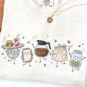 May include: A cream-colored t-shirt featuring a graphic print of five anthropomorphic characters: a serving tray with a burger and drink, a notepad with a pencil, a coffee pot, an apron, and a glass jar filled with cash, accented by small stars and hearts.