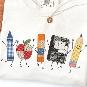 May include: A cream-colored t-shirt featuring a graphic of five anthropomorphic school supplies: a blue gingham crayon, a red patterned apple, an orange glue stick, a black and white composition notebook, and a yellow gingham pencil.
