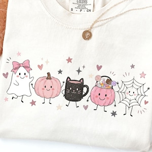 May include: A cream-colored t-shirt featuring a graphic print of a white ghost with a pink bow, a pink pumpkin, a black cat mug, a pink pumpkin bucket filled with candy, and a spiderweb, all with faces, surrounded by small pink and grey stars.