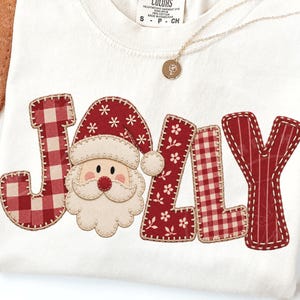 Può includere: Una t-shirt color crema con la parola JOLLY in grandi lettere decorative. La lettera O è sostituita da un volto di Babbo Natale con un cappello rosso a fiocchi di neve bianchi. Le altre lettere presentano motivi a quadri o floreali.