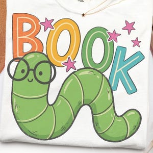 May include: A white t-shirt featuring a graphic of a green worm wearing black round glasses. Above the worm, the word BOOK is printed in large, colorful letters. Several small pink stars are scattered around the text and the worm character.