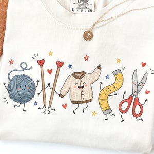 Könnte beinhalten: Ein cremefarbenes T-Shirt mit einem bunten Grafikdruck von anthropomorphen Bastelutensilien, darunter ein blaues Wollknäuel, zwei Stricknadeln mit Herzspitzen, eine beige Strickjacke, ein gelbes Maßband und eine rote Schere.