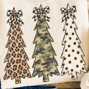 Op de afbeelding: Een crèmekleurig sweatshirt met een grafische print van drie gestileerde kerstbomen. Elke boom is voorzien van een zwart-wit geblokte strik. De eerste boom heeft een luipaardprint, de tweede een camouflagepatroon en de derde is wit met zwarte stippen.