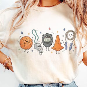 May include: A cream-colored t-shirt featuring a graphic print of five anthropomorphic sports items: an orange basketball, a grey whistle, a black digital stopwatch displaying 00:00, an orange traffic cone, and a jump rope with blue handles.