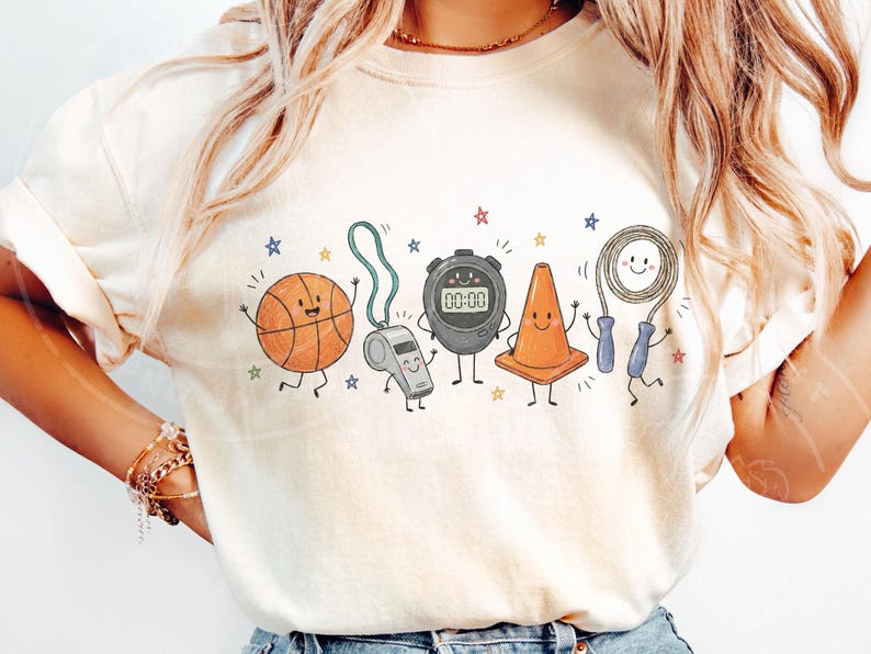 May include: A cream-colored t-shirt featuring a graphic print of five anthropomorphic sports items: an orange basketball, a grey whistle, a black digital stopwatch displaying 00:00, an orange traffic cone, and a jump rope with blue handles.