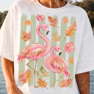 May include: White t-shirt with a design of two pink flamingos and autumn leaves. The design includes green vertical stripes and gold accents. The words "mockup created with AI" are in the upper right corner.