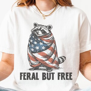 May include: White t-shirt featuring a raccoon wrapped in an American flag. The raccoon illustration is in a vintage style, with the text "FERAL BUT FREE" printed below in a bold font. The shirt is a casual, everyday wear item.