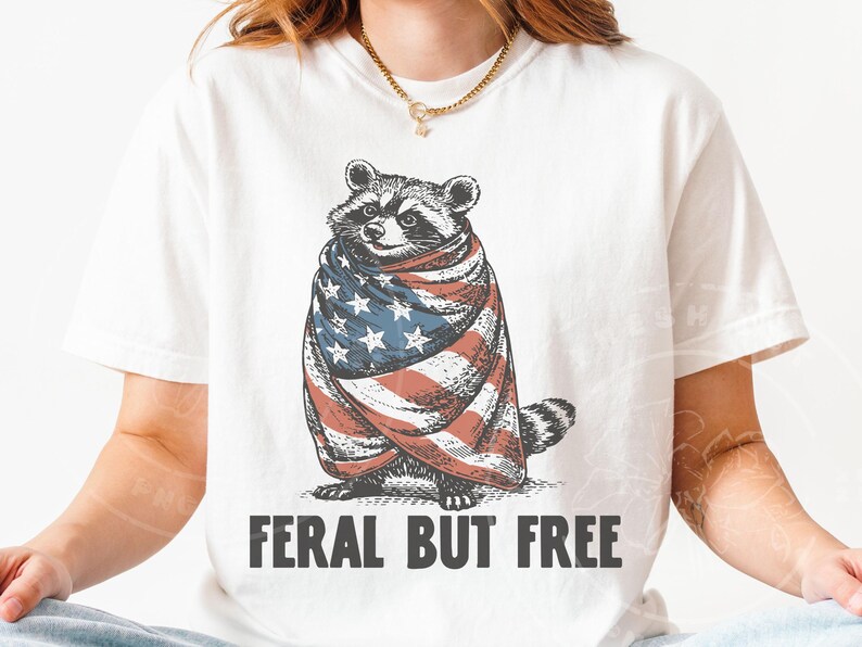 May include: White t-shirt featuring a raccoon wrapped in an American flag. The raccoon illustration is in a vintage style, with the text "FERAL BUT FREE" printed below in a bold font. The shirt is a casual, everyday wear item.