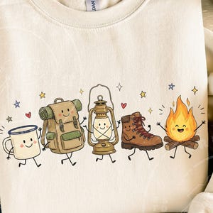 May include: A cream-colored crewneck sweatshirt featuring a colorful graphic print of five anthropomorphic camping items: a white enamel mug, a tan backpack, a gold lantern, a brown hiking boot, and a bright orange campfire, all with smiling faces.