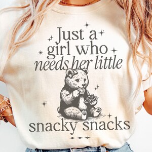 May include: Cream-colored sweatshirt with the text "Just a girl who needs her little snacky snacks." A detailed illustration of a bear holding a flower and a snack is centered below the text. The design is in shades of gray.