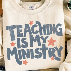 May include: Cream-colored sweatshirt with the text "TEACHING IS MY MINISTRY" in distressed blue lettering. Red stars accent the text. A casual, comfortable top for teachers.