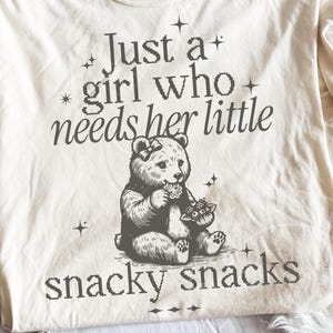 May include: Cream-colored t-shirt with a graphic of a bear eating flowers. The text reads "Just a girl who needs her little snacky snacks" in a pixelated font. The bear has a bow and is surrounded by stars.