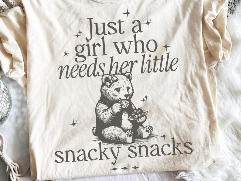 May include: Cream-colored t-shirt with a graphic of a bear eating flowers. The text reads "Just a girl who needs her little snacky snacks" in a pixelated font. The bear has a bow and is surrounded by stars.