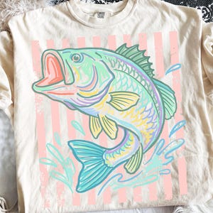 May include: Cream-colored t-shirt featuring a colorful illustration of a leaping fish with an open mouth. The fish is in shades of blue, green, yellow, and purple, set against a backdrop of pink vertical stripes. The shirt has short sleeves.
