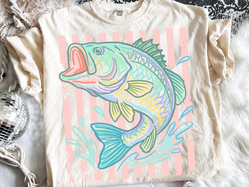 May include: Cream-colored t-shirt featuring a colorful illustration of a leaping fish with an open mouth. The fish is in shades of blue, green, yellow, and purple, set against a backdrop of pink vertical stripes. The shirt has short sleeves.