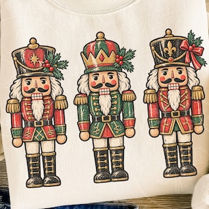May include: A cream-coloured sweatshirt featuring a graphic print of three nutcracker figures. Each nutcracker wears a uniform in shades of red, green, and gold, with black boots and white beards. The figures are arranged side-by-side on the front.