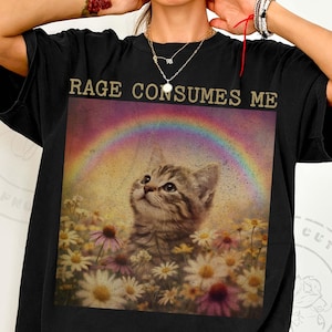 May include: Black t-shirt with a graphic featuring a kitten looking up at a rainbow over a field of daisies. The text "RAGE CONSUMES ME" is printed above the image in gold lettering. The shirt is a casual, oversized style.