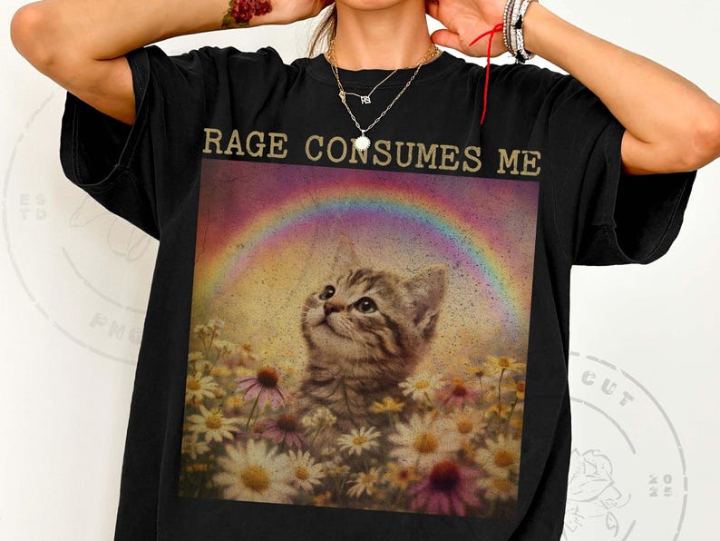 May include: Black t-shirt with a graphic featuring a kitten looking up at a rainbow over a field of daisies. The text "RAGE CONSUMES ME" is printed above the image in gold lettering. The shirt is a casual, oversized style.
