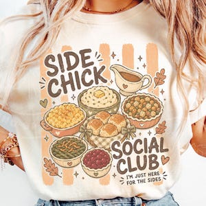 May include: Cream-coloured t-shirt with a graphic featuring various Thanksgiving side dishes and the text "SIDE CHICK SOCIAL CLUB". The design includes a gravy boat, mashed potatoes, stuffing, rolls, and other dishes.