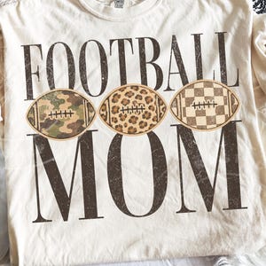 May include: A cream-colored t-shirt featuring the dark brown distressed text FOOTBALL MOM. Between the words are three football graphics: one with a green camouflage pattern, one with a brown leopard print, and one with a brown checkerboard pattern.