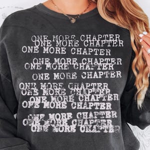 K&ouml;nnte beinhalten: Dunkelgraues Sweatshirt mit dem Text "ONE MORE CHAPTER" in verblichener wei&szlig;er Schrift. Der Schriftzug wiederholt sich mehrfach auf der Vorderseite. Das Sweatshirt hat lange &Auml;rmel und einen Rundhalsausschnitt.