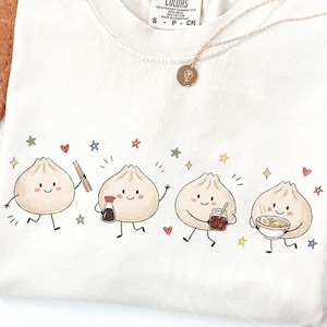 Puede incluir: Una camiseta color crema con un estampado gráfico de cuatro dumplings animados con caras, brazos y piernas. Los dumplings interactúan con palillos, una botella de salsa de soja, un frasco de aceite de chile y un tazón de ramen, rodeados de estrellas.