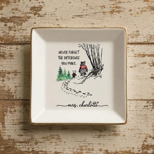 May include: A square white ceramic trinket dish with a gold-toned rim. The center features a black ink illustration of two bears walking away, green pine trees, and the text: NEVER FORGET THE DIFFERENCE YOU MAKE. Below is the name mrs. charlotte.
