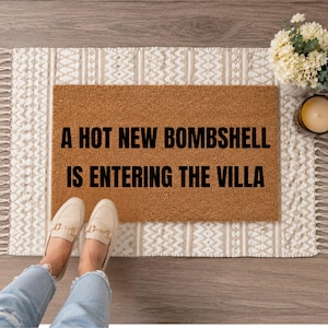 May include: A rectangular brown doormat with the text "A HOT NEW BOMBSHELL IS ENTERING THE VILLA" in black. The doormat is on a patterned rug with a cream and beige design. A pair of beige loafers with gold accents are visible.