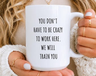 May include: A white ceramic mug featuring black printed text that reads: YOU DON'T HAVE TO BE CRAZY TO WORK HERE. WE WILL TRAIN YOU. The mug has a standard handle and a smooth, glossy finish, displaying the message clearly in a simple sans-serif font.