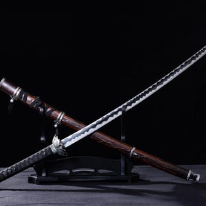 May include: A Japanese katana sword with a dark wooden scabbard and handle, displayed on a black stand. The blade is silver with a black wavy pattern. The handle is wrapped in black fabric.