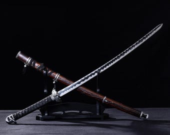 May include: A Japanese katana sword with a dark wooden scabbard and handle, displayed on a black stand. The blade is silver with a black wavy pattern. The handle is wrapped in black fabric.