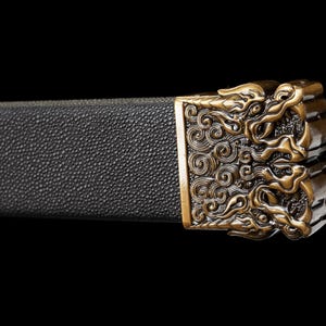 May include: Close-up of a sword hilt with a black textured grip and an ornate, gold-colored metal guard. The guard features a detailed design of swirling patterns and stylized figures, set against a black background.