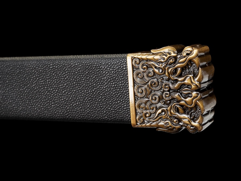 May include: Close-up of a sword hilt with a black textured grip and an ornate, gold-colored metal guard. The guard features a detailed design of swirling patterns and stylized figures, set against a black background.