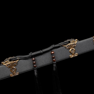 May include: Close-up of a black sword scabbard with ornate gold-colored metal accents. The scabbard features decorative clasps and a detailed end piece. Black tassels and a braided cord add to the design.