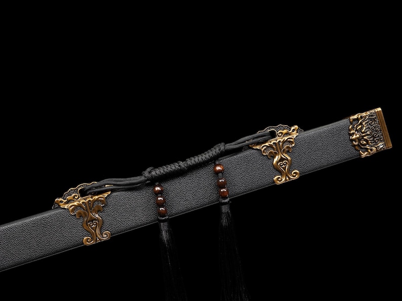 May include: Close-up of a black sword scabbard with ornate gold-colored metal accents. The scabbard features decorative clasps and a detailed end piece. Black tassels and a braided cord add to the design.