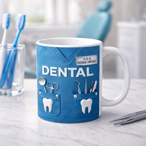 May include: White ceramic mug with a blue dental scrubs design. The mug features the word "DENTAL" in white block letters, along with a name tag that reads "D.D.S. Honda Hiroaki". The design includes pockets with dental tools and tooth graphics.