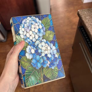 May include: A rectangular book with a cover featuring a stained glass style illustration of blue and white hydrangea flowers with green leaves. The design uses dark blue outlines to separate the colored sections, creating a mosaic appearance.
