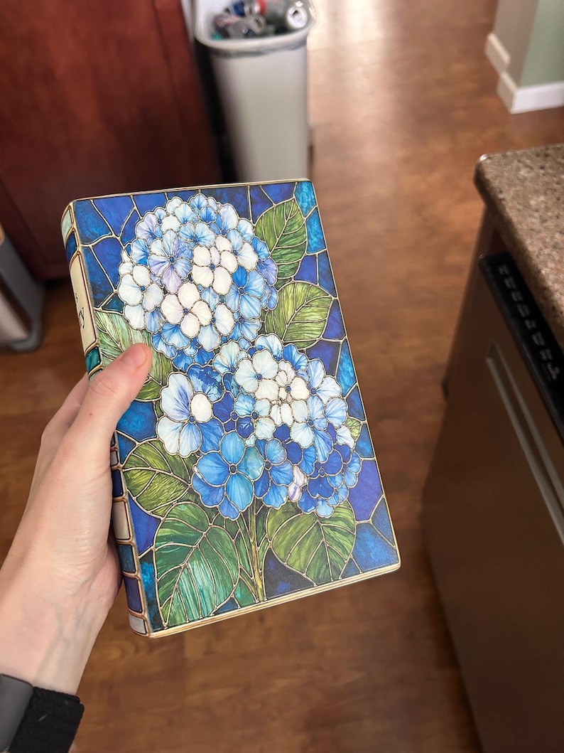 May include: A rectangular book with a cover featuring a stained glass style illustration of blue and white hydrangea flowers with green leaves. The design uses dark blue outlines to separate the colored sections, creating a mosaic appearance.