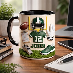 Puede incluir: Taza de cerámica con asa y borde negros, decorada con elementos de fútbol americano. El diseño incluye un casco, una camiseta con el número 12, un balón de fútbol y una portería. El nombre "JOHN" está presente.