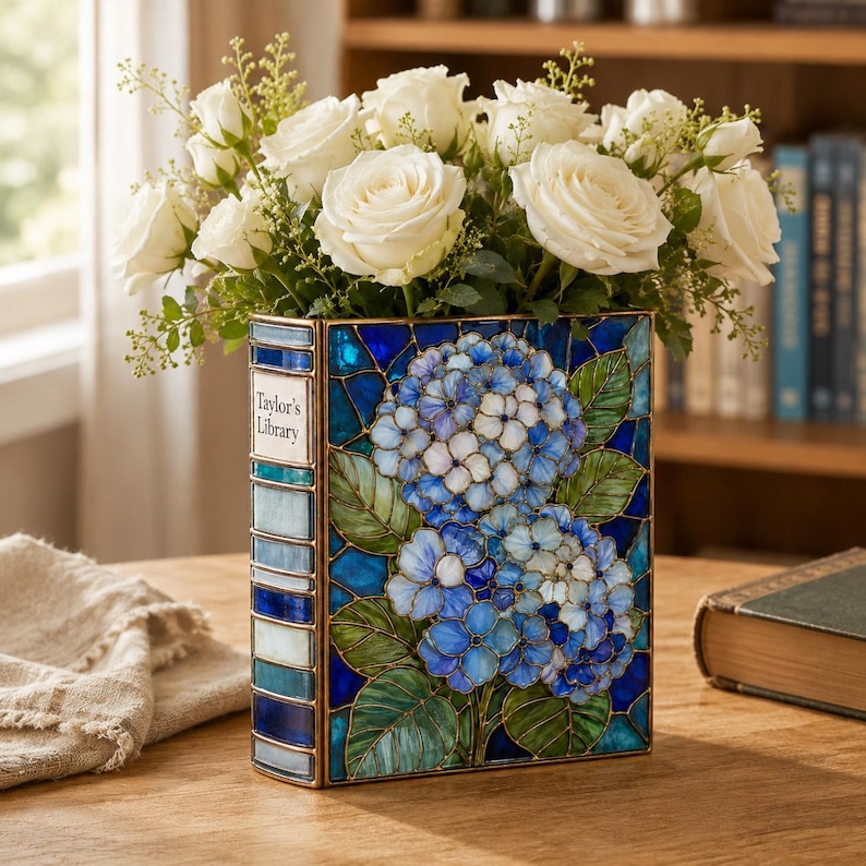May include: A rectangular stained glass vase shaped like a book with a blue and green hydrangea floral pattern and the text Taylor's Library on the spine. The vase is filled with a bouquet of white roses and small green filler flowers.