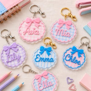 May include: Six circular acrylic keychains with scalloped edges, each featuring a pastel gingham pattern, a raised bow, and a personalised name in script font. The keychains include silver or gold metal lobster clasps and split rings.
