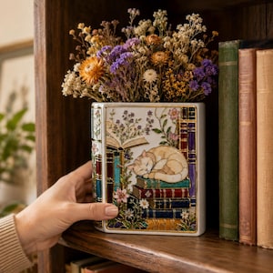 May include: A white ceramic book-shaped vase featuring a colorful illustration of a sleeping cat curled atop a stack of books, surrounded by floral accents and an open book design. The vase is filled with a bouquet of dried orange, purple, and white flowers.