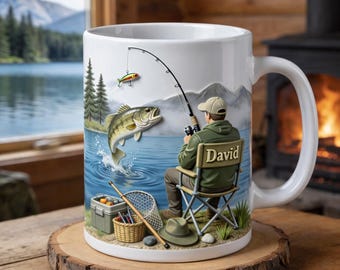 Puede incluir: Taza de cerámica blanca con una escena de pesca. El diseño presenta un pescador, un pez saltando y aparejos de pesca. La silla del pescador tiene el nombre "David" impreso. El fondo muestra un lago, montañas y árboles.