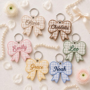 May include: Six embroidered bow-shaped keychains featuring gingham patterns in beige, pink, yellow, green, and blue. Each keychain is personalised with a name stitched in matching thread and includes a silver metal ring for attachment.