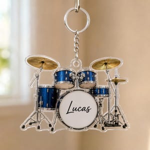 Peut inclure: Un porte-clés en acrylique transparent en forme de batterie bleue avec des cymbales dorées et des pieds argentés. La grosse caisse porte le prénom Lucas écrit en noir. L'objet est suspendu à un anneau métallique et une chaînette.