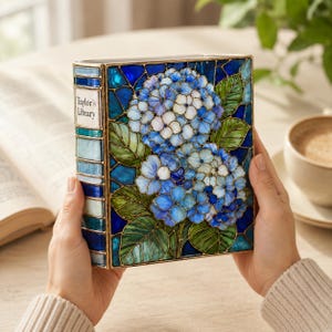 May include: A rectangular decorative box designed to resemble a book with a stained glass style cover featuring blue and white hydrangea flowers and green leaves. The spine is labeled Taylor's Library in black text on a white background.