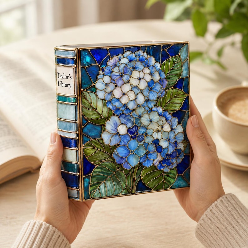 May include: A rectangular decorative box designed to resemble a book with a stained glass style cover featuring blue and white hydrangea flowers and green leaves. The spine is labeled Taylor's Library in black text on a white background.