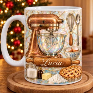 May include: A white ceramic mug featuring a copper-colored stand mixer, a whisk, a spoon, a bowl with eggshells, a rolling pin with the name Lucia, a jar of sugar, a bottle of extract, a stick of butter, flour, and a fruit pie.