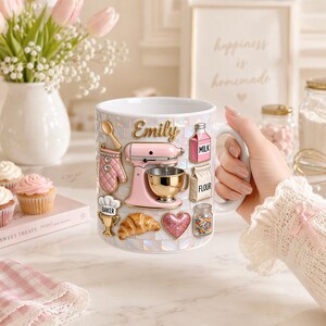 May include: A white ceramic mug with a mosaic tile pattern, adorned with baking-themed embellishments. Features a pink stand mixer, a croissant, and the name "Emily". Includes a jar of sprinkles and a milk carton.